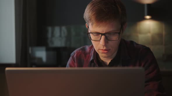 A Man in Glasses Works at a Laptop in a Home Interior in the Evening