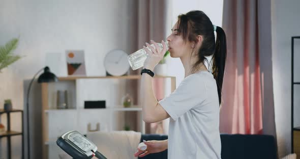 Woman with Ponytail Drinking Water During Training on Treadmill at Home alt