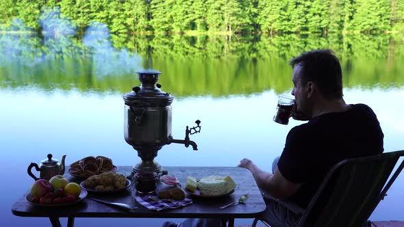 Man drinking tea by the lake. Vintage tea samovar with smoke and food near lake alt