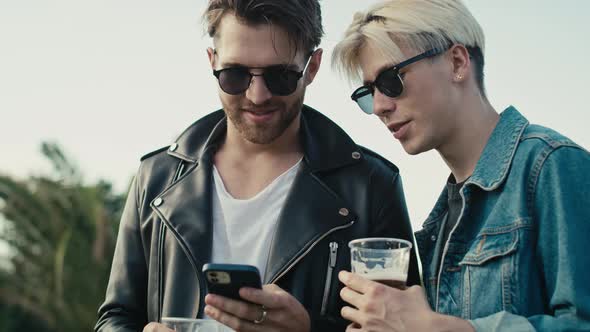 Two young caucasian men browsing phone and having fun on music festival while drinking beer. Shot wi alt