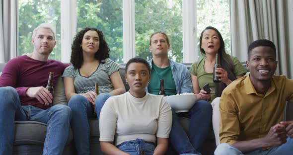 Diverse group of happy male and female friends watching sport drinking beer, cheering in living room alt