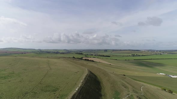Aerial tracking slowly west above the northern ramparts of Maiden Castle. Beautiful green fields and alt