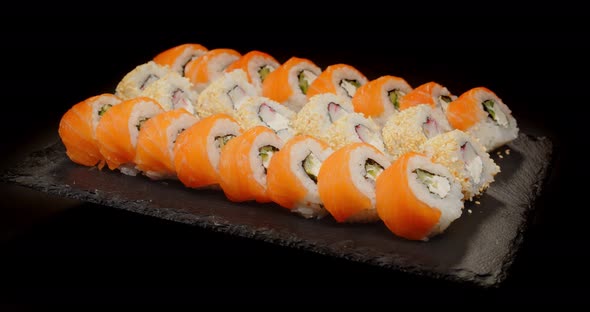 Sushi Wrapped In Salmon And Sprinkled With Sesame Seeds Are Laid Out On A Black Board
