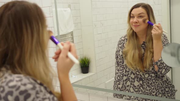 Beautiful Young Woman Doing Makeup with a Big Fluffy Brush in the Bath in Front of a Mirror alt