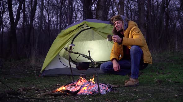 A Man Drinks Tea Near a Campfire and a Tent in the Forest alt