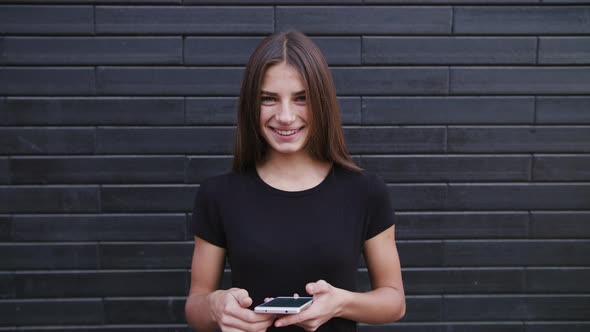An Attractive Young Lady Wearing Glasses Using a Phone Against a Brick Wall Background. Close-up alt