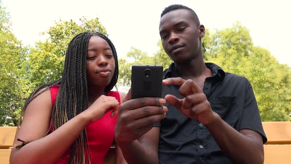 A Black Woman Teaches a Black Man Something on a Smartphone As They Sit on a Bench alt