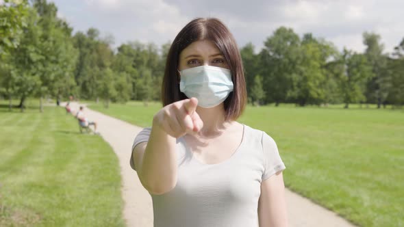 A Young Caucasian Woman in a Face Mask Points at the Camera and Nods on a Pathway in a Park alt