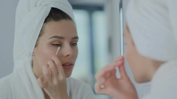 Focused brunette woman removing make up with cotton stick and looking at reflection in mirror alt