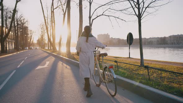 Woman in Beige Coat Carries Yellow Bicycle Along Road alt