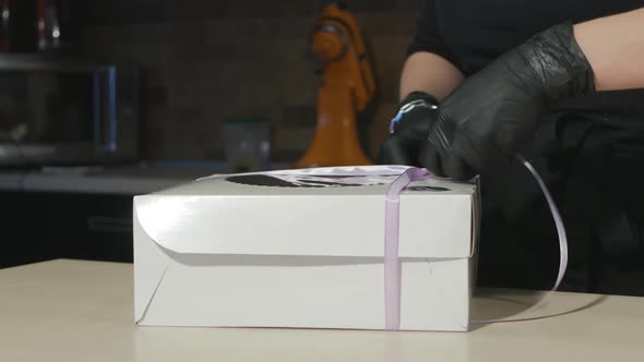 the Hands of a Woman Pastry Chef in Black Gloves Tie a Ribbon with a Bow on a Box with a Dessert