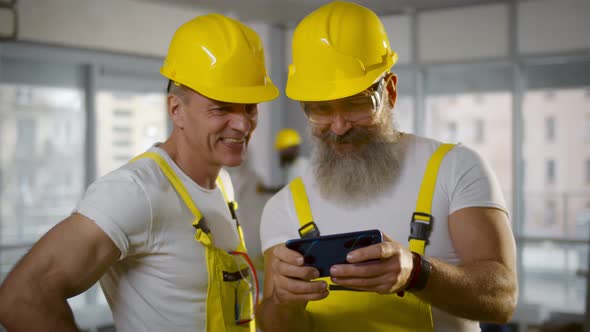 Two Builders in Hardhat and Overall Using Smartphone at Construction Site alt