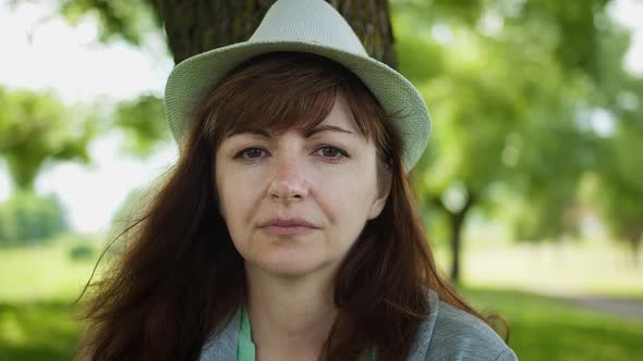 Portrait of a Woman in a Hat Near the Tree in the Park Looking at the Camera