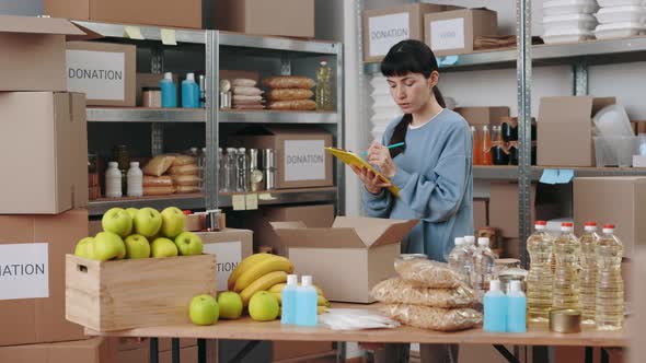 Woman Writing on Clipboard Among Food Bank Warehouse alt