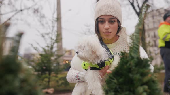Woman with a White Dog in Her Arms Near a Green Christmas Trees alt