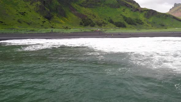 Waves crashing on black sand beach in Vik, Iceland with drone over water moving forward. alt