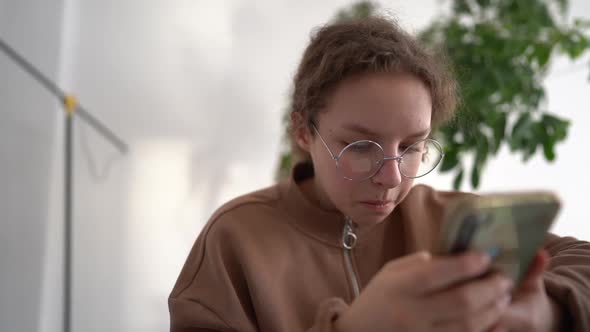 Teen Girl Consulting with Her Friend Using Modern Phone While Sitting at Desk Table in Living Room alt