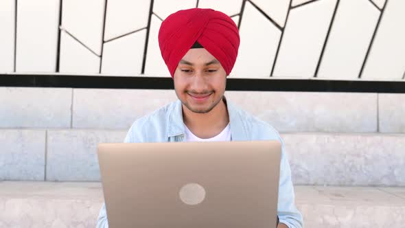 Cheerful Indian Man Wearing Headwrap Turban Sitting on the Steps Outdoors and Using Laptop