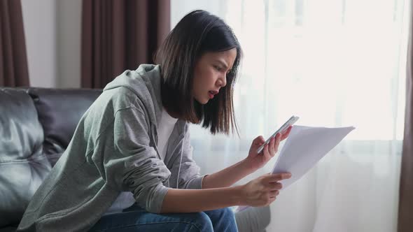 Asian woman holding the bill and smartphone in the living room and stressed by excessive expenses. alt
