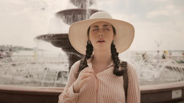 Sweaty woman in a straw hat is standing on the street, fanning herself with her hand from the heat alt