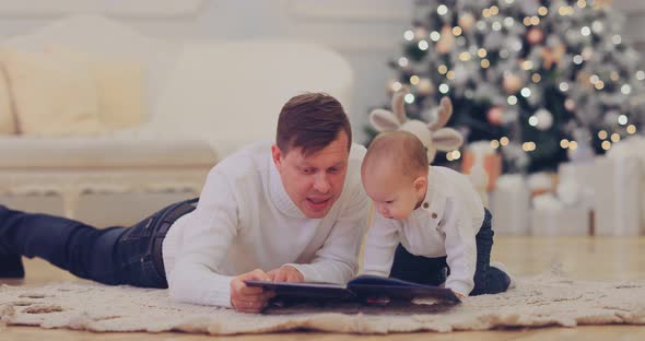 Happy Dad with His Son Lie By the Beautiful New Year Tree and Read a Book with Fairy Tales alt