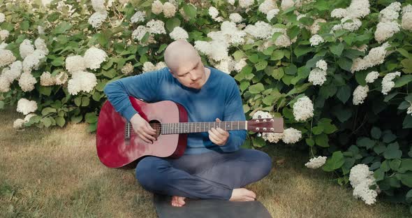 Man with an Acoustic Guitar is Sitting in a Park in Nature and Tuning Instrument