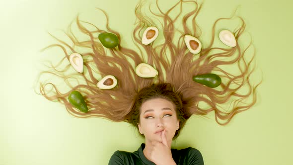 beautiful girl lying on green background with avocado fruits on long hair, woman thinking,choice  alt