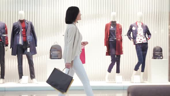 Young Woman Walks Along a Show Window with Bags and Phone in Her Hand in the Shopping Mall alt