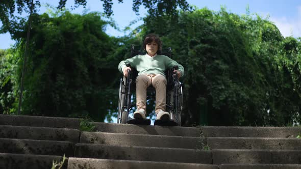 Front View Portrait Sad Disabled Boy in Wheelchair at Stairs with Summer Park at Background alt