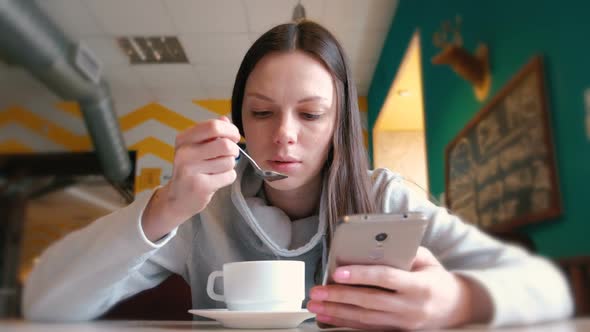 Woman Is Drinking a Hot Chocolate with Spoon and Look on the Screen of Mobile Phone