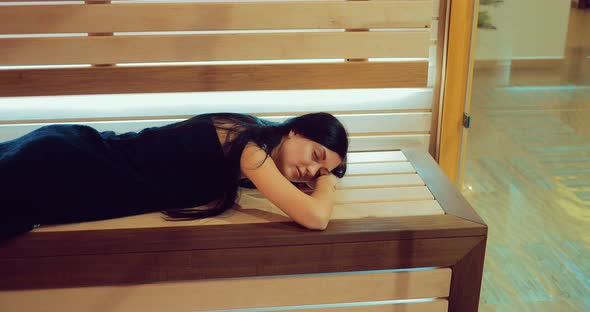 Beautiful Brunette Girl is Lying on a Wooden Bench in a Sauna Behind a Glass Door alt