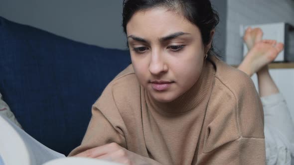 Close Up Of A Young Indian Woman Lying On A Bed Reading A Book Her Hand Turns The Page alt
