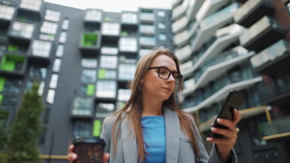 Formally Dressed Woman Walking Down the Street in a Business District with Coffee in Hand and Using alt