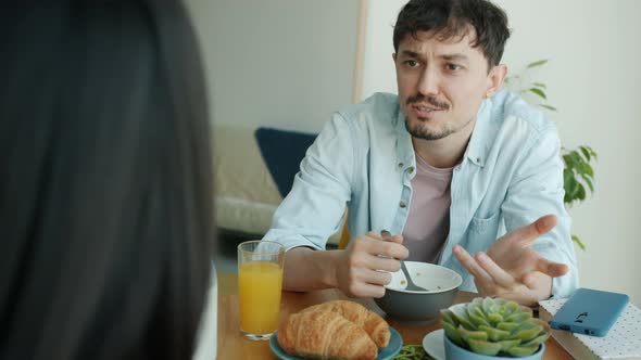 Happy Young Man Eating Breakfast and Talking to Woman Sitting at Table in Kitchen alt
