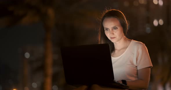 Young Female Student with Laptop at Night in Town Typing Text alt