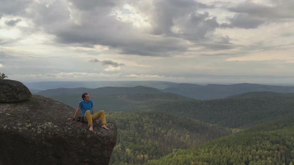 Aerial Shot of a Young Man Sitting on the Edge of a Cliff Enjoying the Solitude