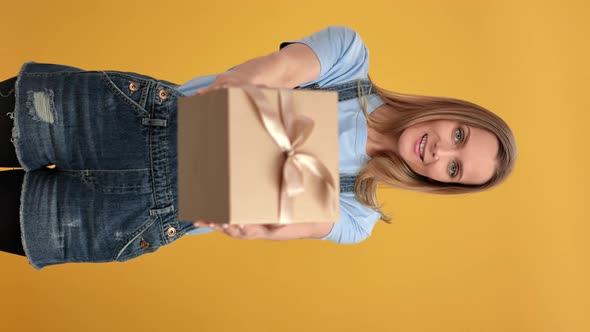 VERTICAL VIDEO POV Blonde Woman Giving Craft Wrapped Gift Box with Bow Posing Isolated on Orange alt