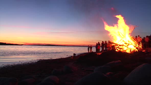 A big bonfire party during a sunset on a beach by the ocean. Kids playing around the fire. alt