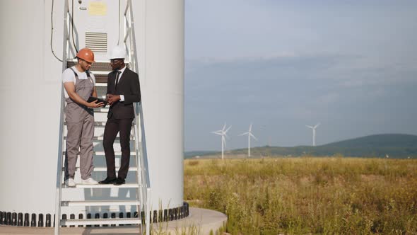 Multiracial Partners Standing on Stairs Near Wind Turbine and Looking on Screen alt