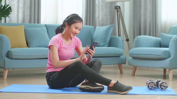 Asian Female Fitness Girl Is Using An App On Her Smartphone Before Working Out In Living Room alt