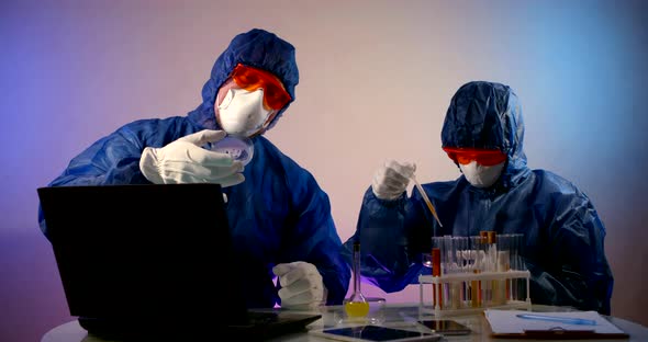 Two People in Protective Clothing, Respirators, Glasses and Gloves Are Sitting at a Table with a alt