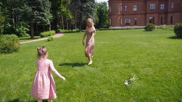 Woman and a Child in Pink Dresses Run Across a Green Field Against the Background of an Old Castle alt