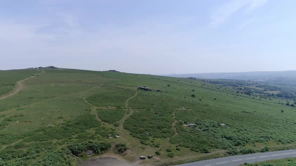 Wide aerial tracking forward over the vast wilderness of Dartmoor, crossing over a road and carpark. alt