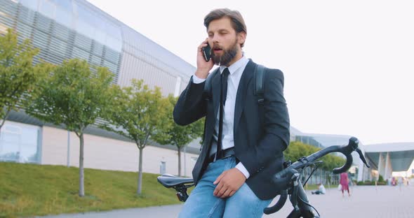 Bearded Man in Stylish Clothes Standing Near His Bike and Talking on Phone alt