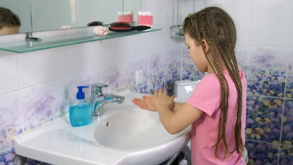 A little girl washes her hands properly with soap and running water over the white sink 