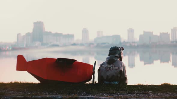 Back View of Little Pilot Boy Sitting Near Amazing Peaceful City Lake alt