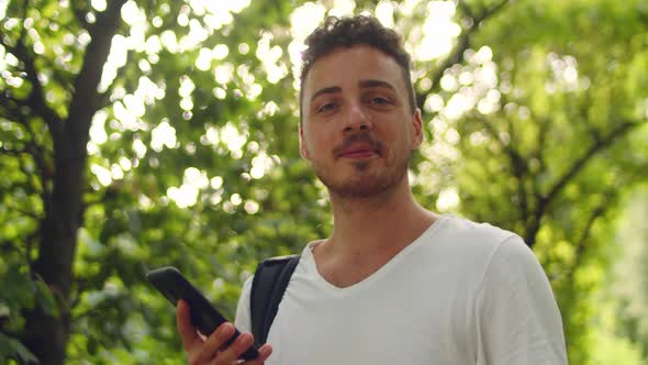 Handsome Young Man Stands in the Park Inside a Tunnel of Trees with a Phone in His Hands alt
