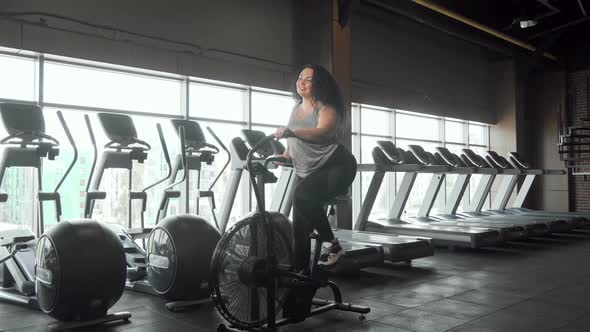 Full Length Shot of a Happy Plus Size Woman Working Out on Air Bike at the Gym alt