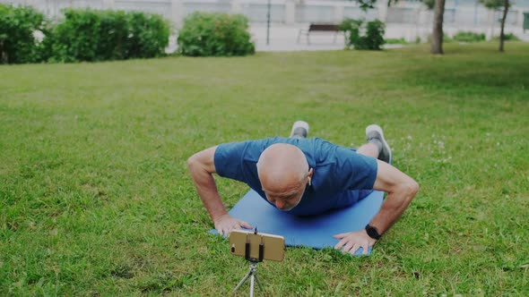 Powerful Old Male Athlete Makes Push Up While Looking on Smartphone Screen alt