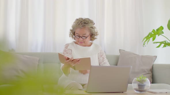 Asian senior woman hold book and learning with computer laptop sit on sofa at home alt